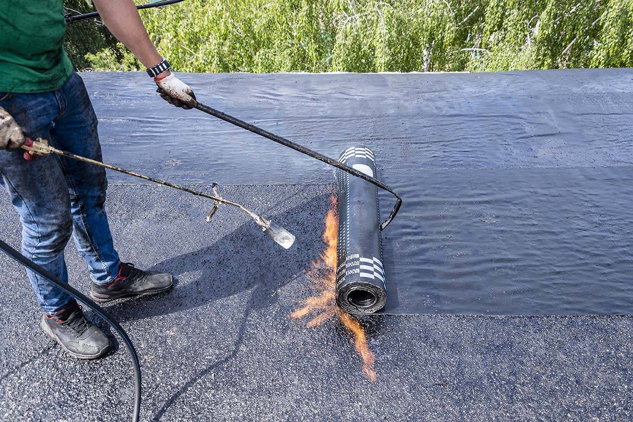 A worker rolls out black roofing material while heating it with a propane torch, surrounded by greenery, on a sunny day.