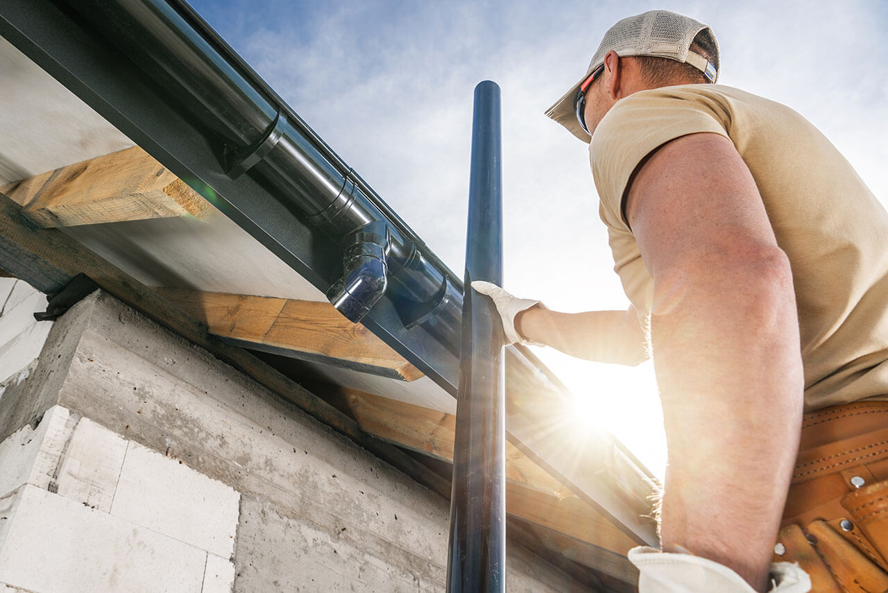 A construction worker attaches a pipe to a roof, sunlight shining in the background, with wooden beams and concrete visible.