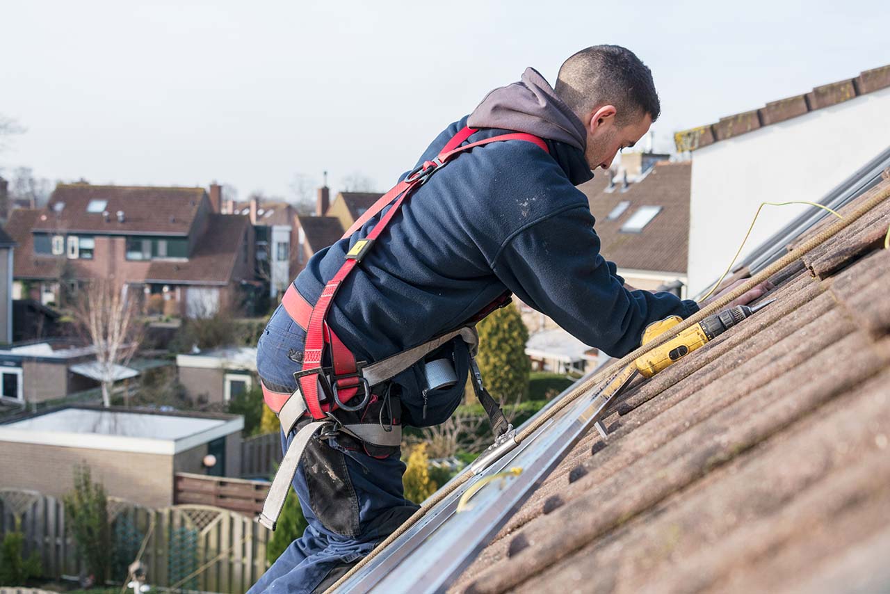 A man in a harness works on a sloped roof, using a drill to install solar panels, with residential houses in the background.