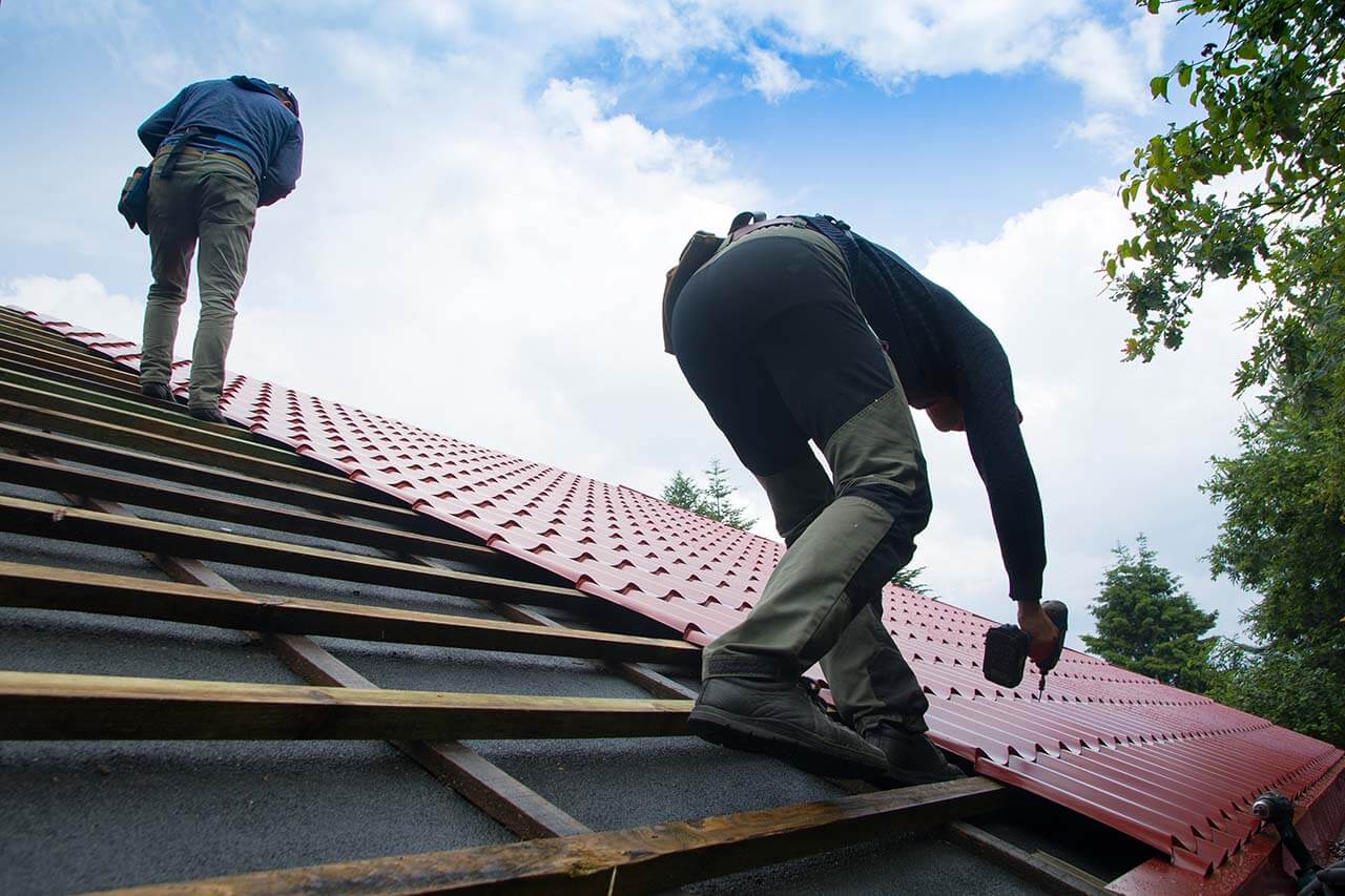 Two workers install red metal roofing on a sloped structure, with one using a power drill and the other inspecting from above.