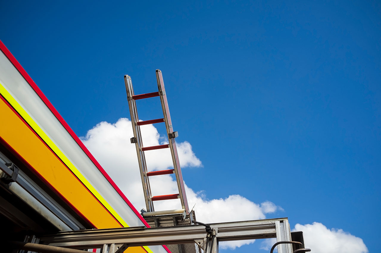 A tall ladder extends upwards against a bright blue sky, with fluffy clouds and the colorful edge of a building visible below.