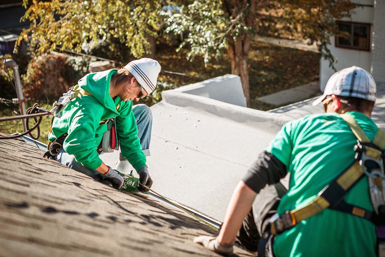 Two workers in green shirts and safety gear are installing materials on a sloped roof, surrounded by autumn foliage.