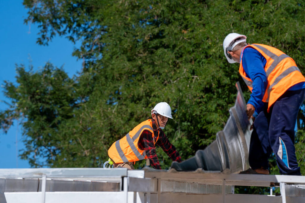 Two workers in safety gear are installing roofing materials on a structure under a clear blue sky, surrounded by greenery.